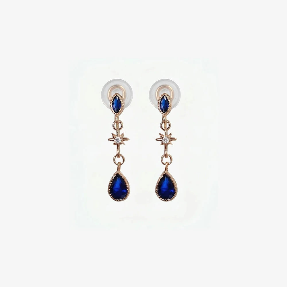 A pair of rose gold clip-on earrings featuring a large navy blue teardrop sapphire and a sparkling crystal starburst accent on a clean white background.