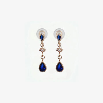 A pair of rose gold clip-on earrings featuring a large navy blue teardrop sapphire and a sparkling crystal starburst accent on a clean white background.