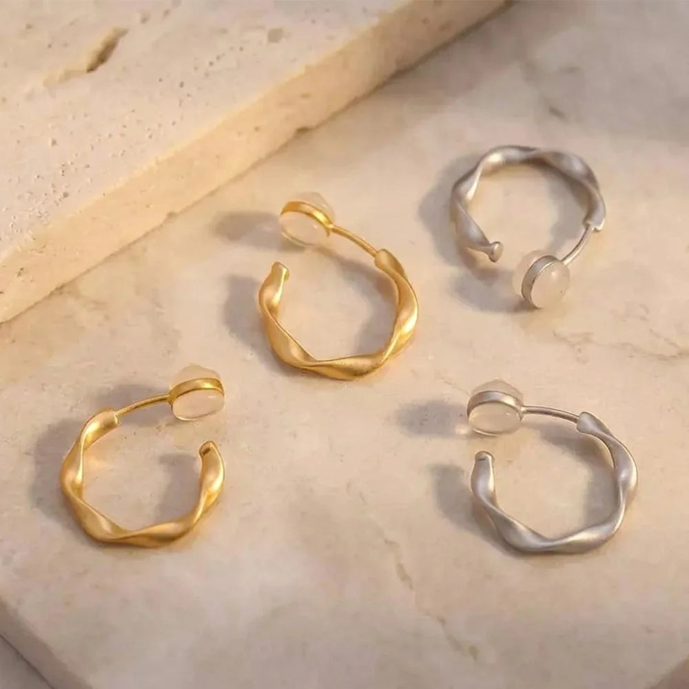 A pair of matte gold sculptural clip on hoop earrings and a pair of matte white gold sculptural clip on hoop earrings shown together on marble surface.