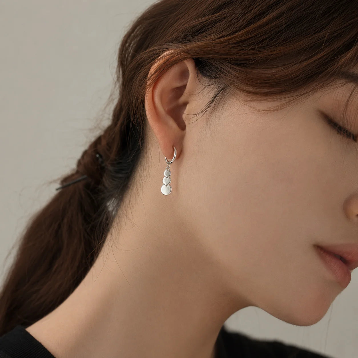 A close-up of a woman's ear wearing a delicate silver huggie hoop earring with three small stacked silver disc charms.