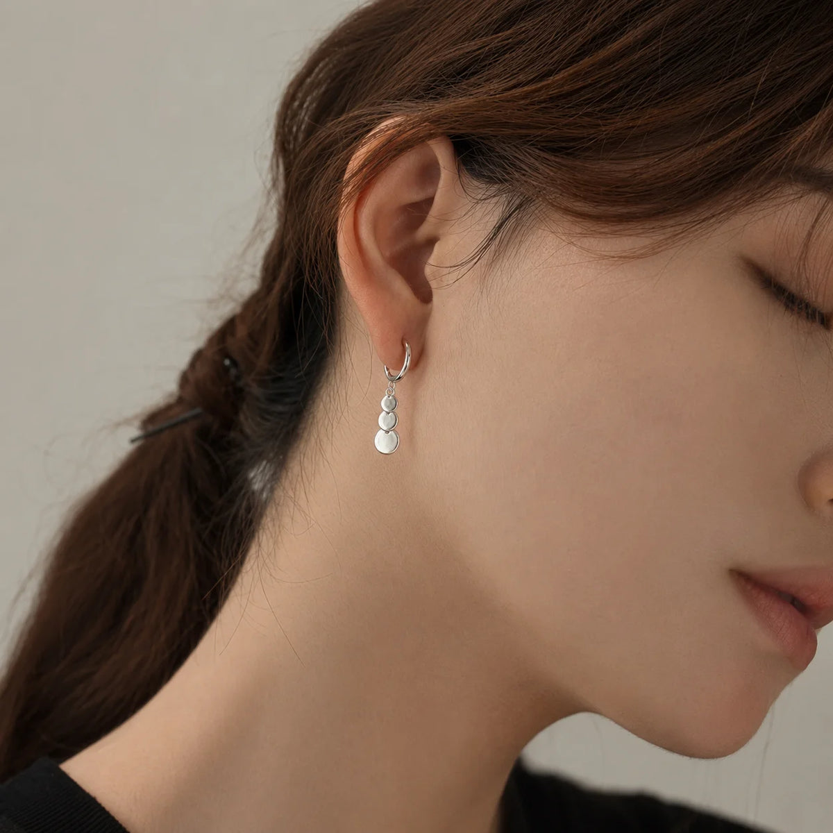 A close-up of a woman's ear wearing a delicate silver huggie hoop earring with three small stacked silver disc charms.