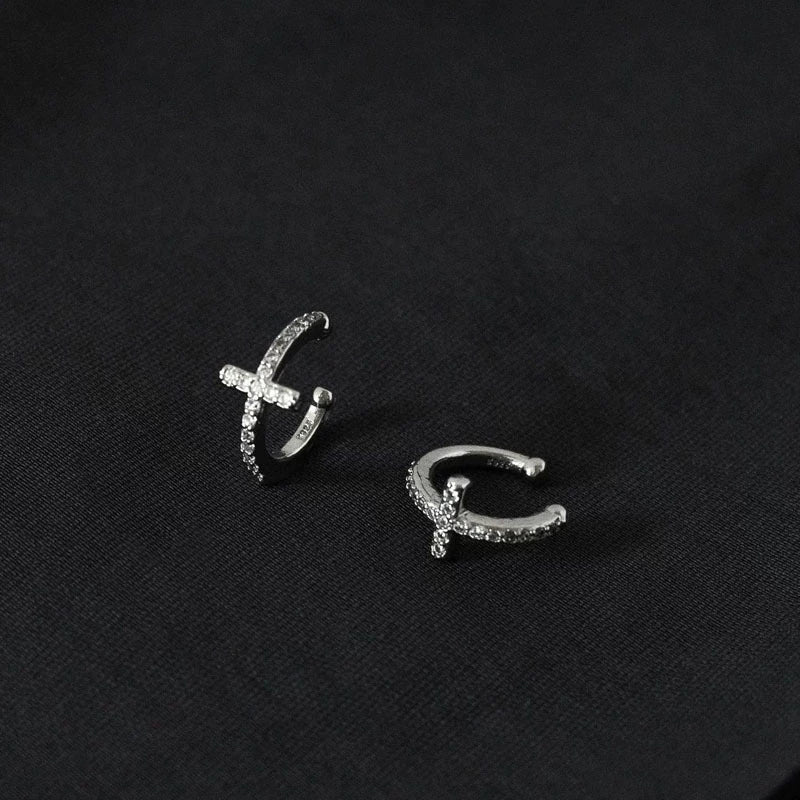 A pair of silver pavé cross clip-on ear cuffs, elegantly arranged on a dark, textured background to highlight their shine.