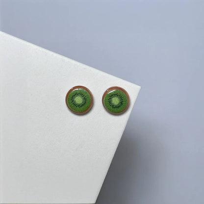A pair of Sweet Kiwi Magnetic Stud Earrings, featuring realistic green kiwi slice designs, resting on a white card at an angle.
