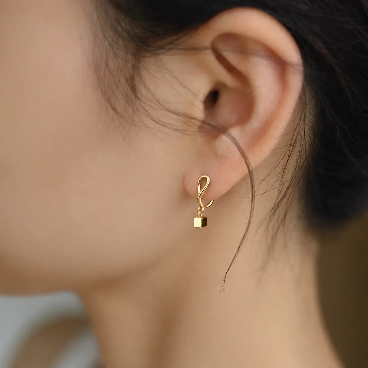 Tiny gold cube charm clip-on earring worn on a non-pierced earlobe, looking pierced from the front