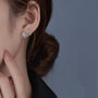 A close-up of a woman's ear wearing a pair of silver star-shaped stud earrings with light blue and orange gemstones, set against a dark gray background.