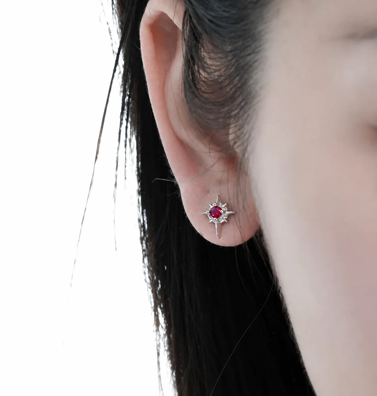 A close-up of a person's ear wearing a small silver-toned starburst stud earring with a red gemstone center.