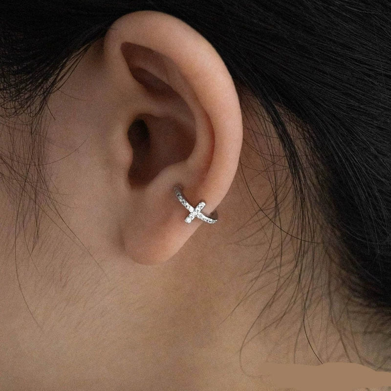 A close-up of a woman's ear wearing a single silver pavé cross clip-on ear cuff on her lobe, showing its delicate sparkle.