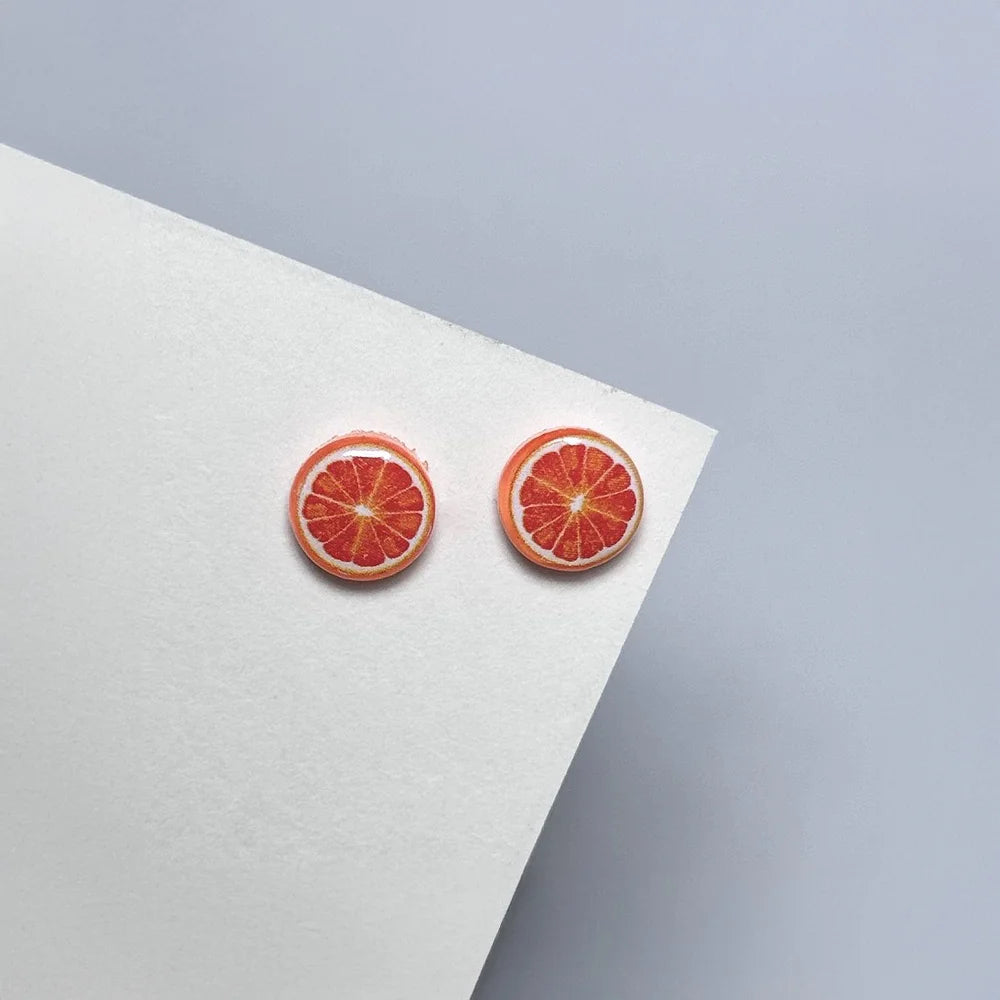 A pair of Zesty Grapefruit Magnetic Stud Earrings, featuring realistic fruit slice designs, resting on a white card at an angle, highlighting the resin texture.