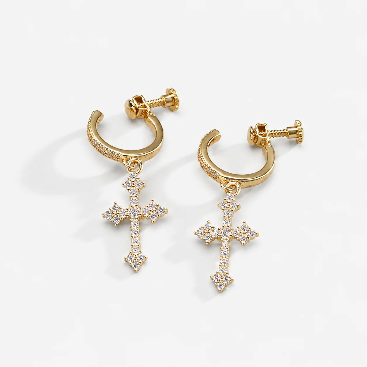 A pair of gold huggie hoop earrings with dangling cross charms, embellished with small clear pavé stones, shown on a clean white background.